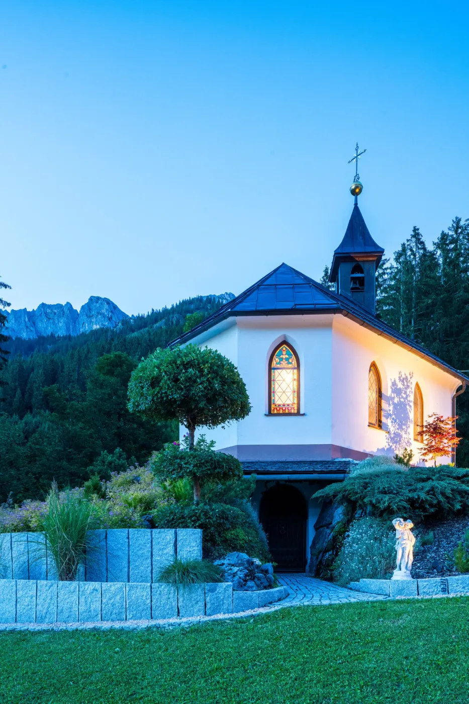 Die Kapelle Sankt Antonius in der blauen Stunde, mit beleuchteten Fenstern vor den Tannheimer Bergen