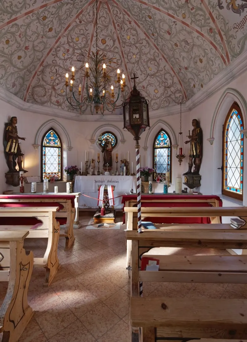Der Innenraum der Kapelle St. Antonius mit Holzbänken, Kerzenleuchter, bemalter Decke und farbigen Bleiglasfenstern